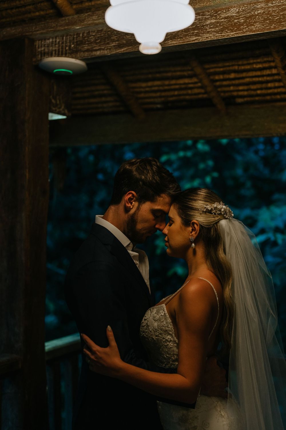 A romantic wedding couple shares an intimate moment under wooden beams in moody lighting.