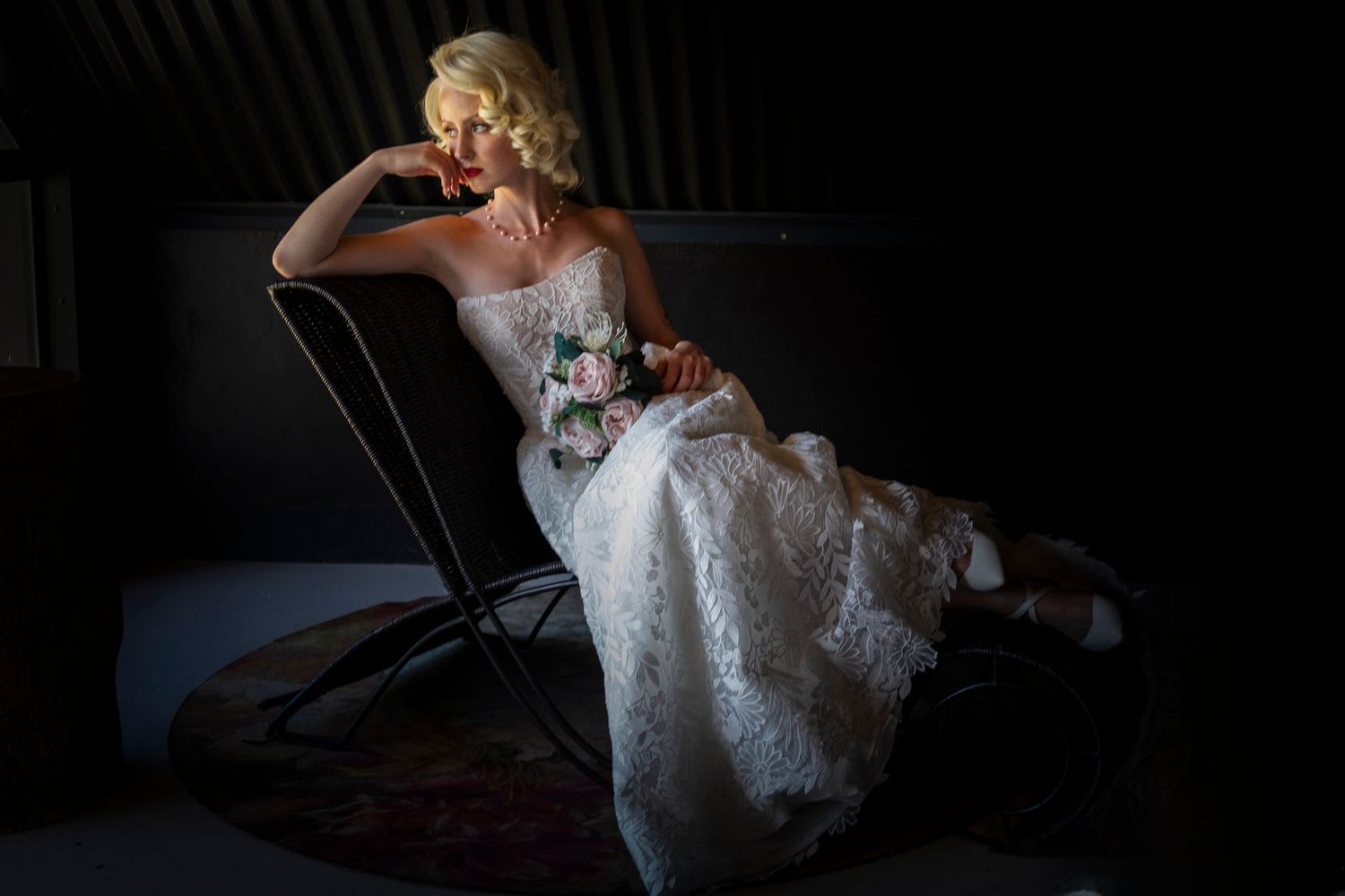 Elegant white lace wedding dress illuminated in dramatic lighting while posed on vintage chair against dark background.