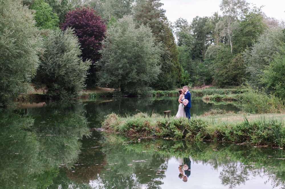 Wedding couple poses by a serene pond surrounded by lush greenery and trees.