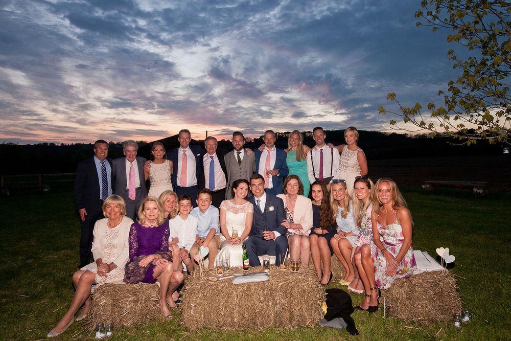 Wedding party poses together on rustic hay bales at sunset during outdoor country celebration.