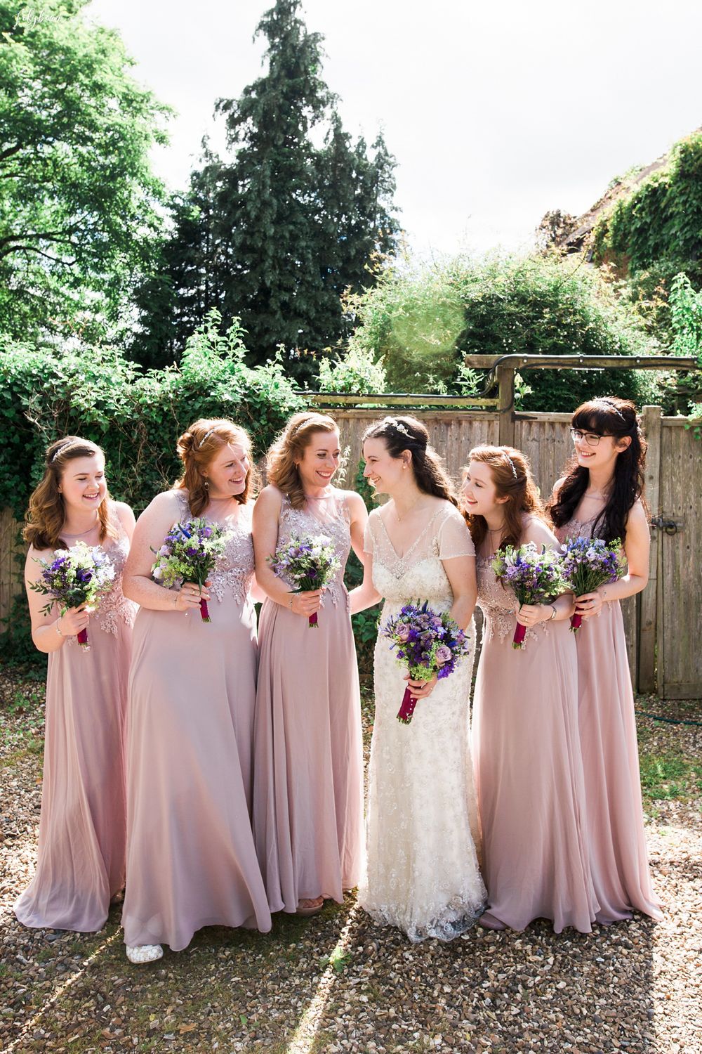 Bridesmaids in flowing blush pink dresses share candid moments while holding bouquets in an outdoor garden setting.