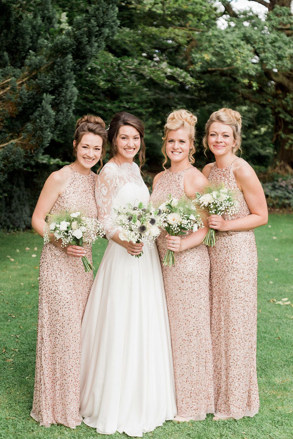Bridal party in rose gold sequin dresses holding white floral bouquets on green lawn.