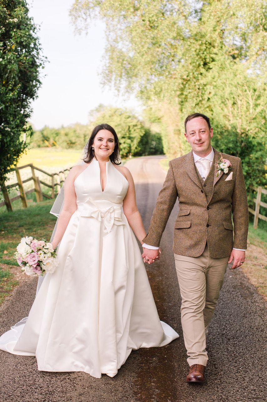 Couple walks hand in hand down tree-lined path in wedding attire.