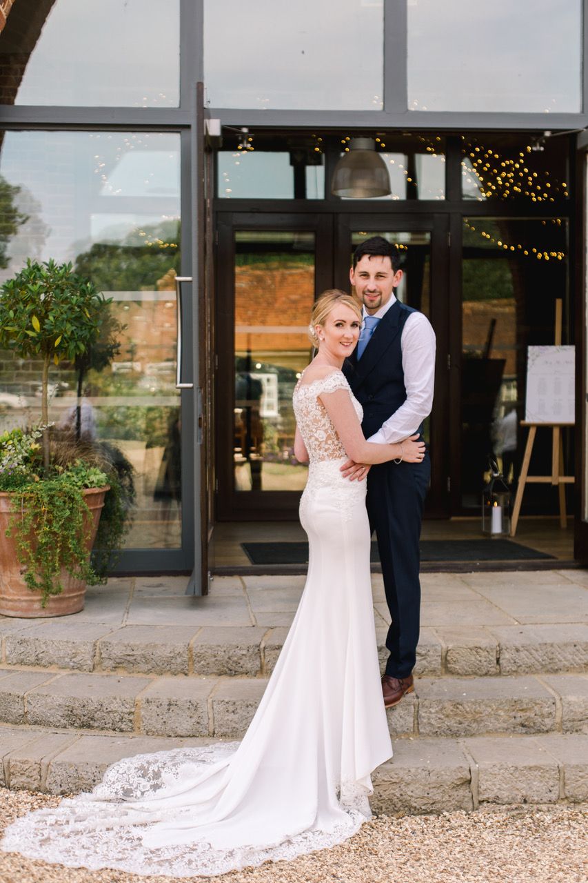 Wedding couple embraces outside brick venue with stunning lace train detail.