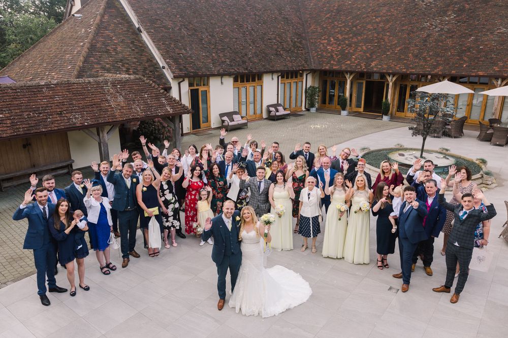 Large wedding party and guests gathered outside traditional brick building venue.
