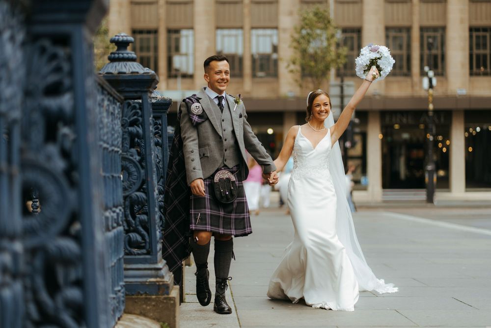 Bride and groom walk through a city street with ornate buildings and lamp posts in the background.