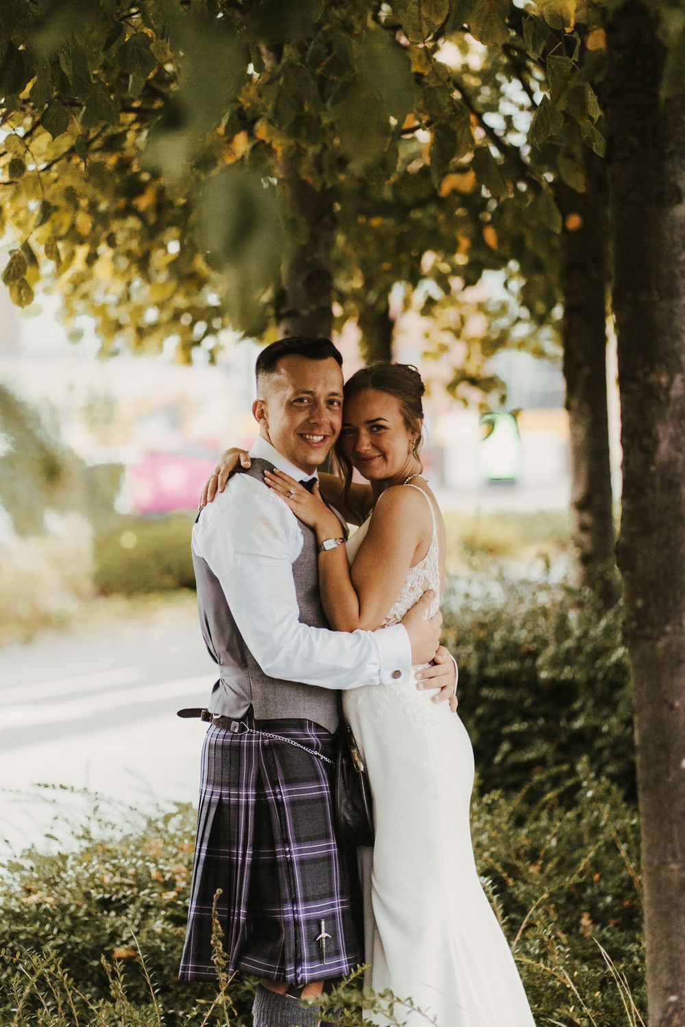 A bride and groom embrace outdoors, with the groom wearing a traditional Scottish kilt and the bride in a fitted white gown.