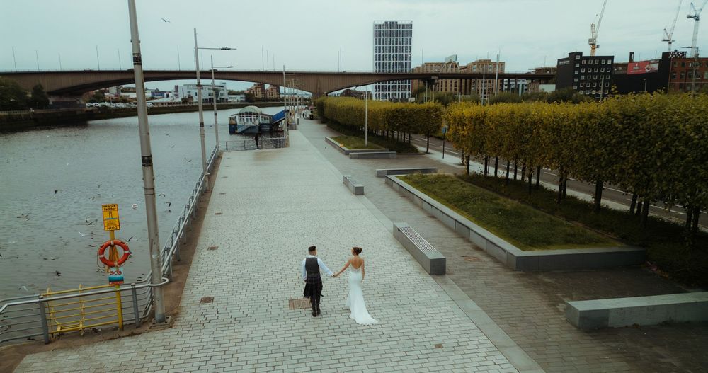 Two figures walk along a waterfront promenade near a harbor with boats and buildings in the background.