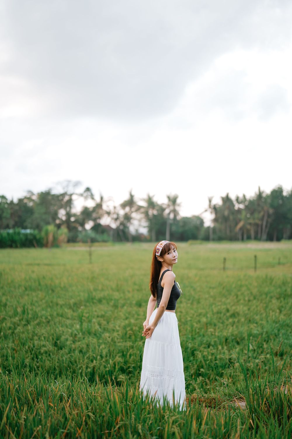 Person in white dress walking through lush green field with palm trees in distance.