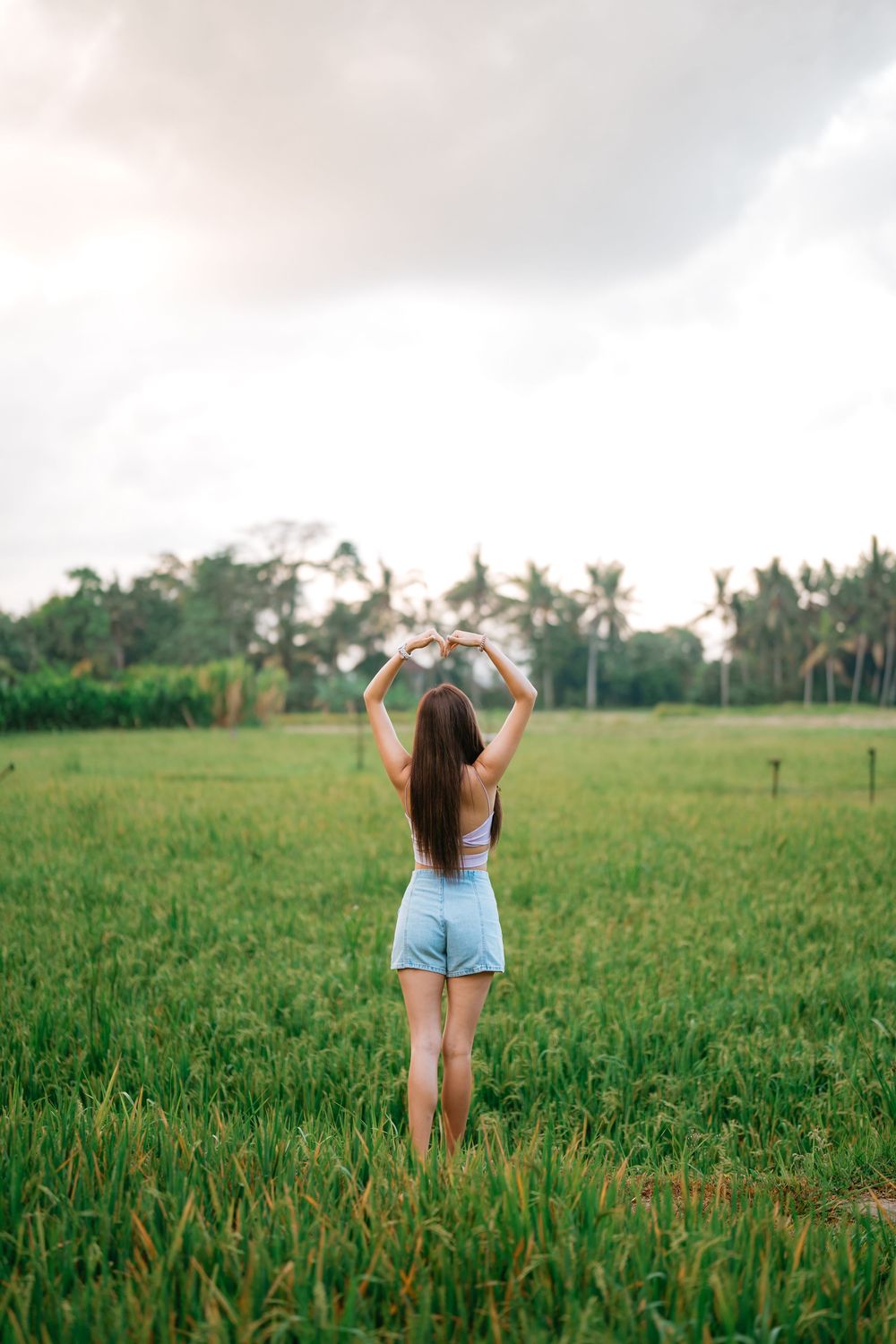 Person with raised arms enjoying moment in sunlit green field with palm trees.