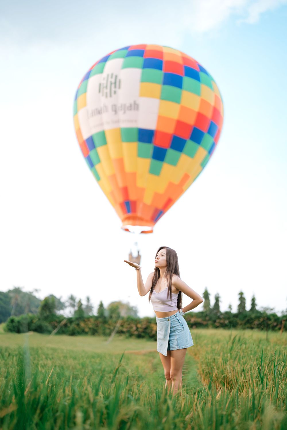 Multicolored hot air balloon drifting over a summer field with trees in the background.