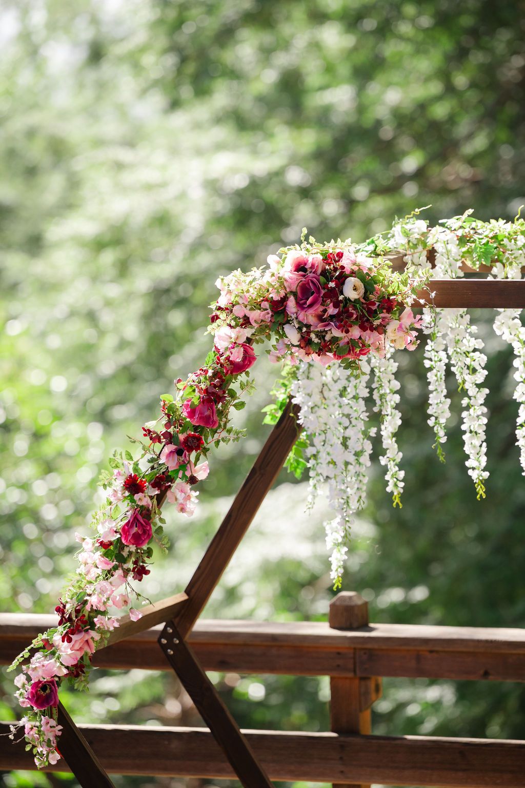 Decorative floral arch with pink and white blooms adorns a wooden pergola structure in a serene forest setting.
