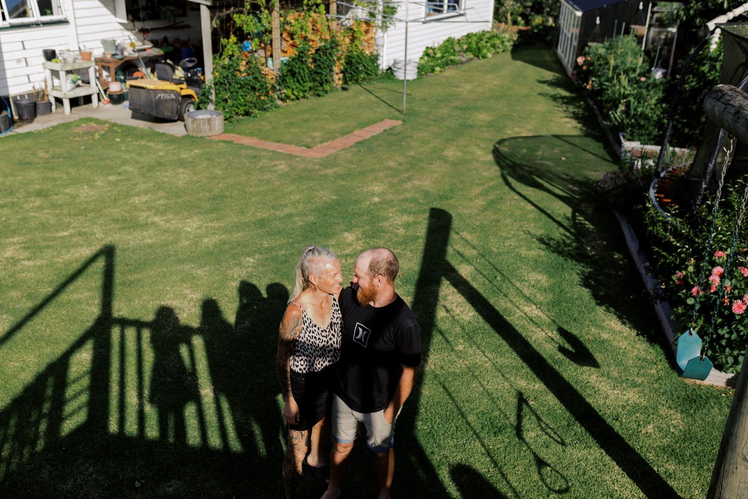 People gather on a sunny lawn near flowering plants and garden furniture in a backyard setting.