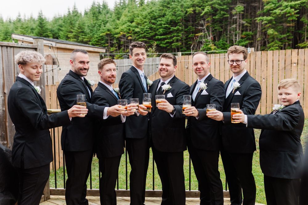 Groomsmen in black tuxedos toast with drinks while standing against a wooden fence outdoors near pine trees.