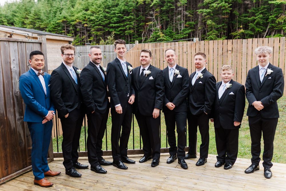 Group of groomsmen in black suits pose sitting on wooden benches and leaning against rustic fence outdoors.