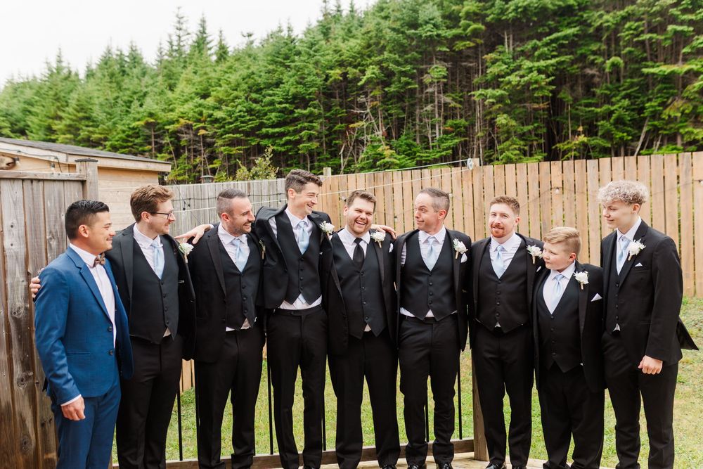 Wedding party in formal attire gather for a group photo against a rustic wooden fence with evergreen forest backdrop.