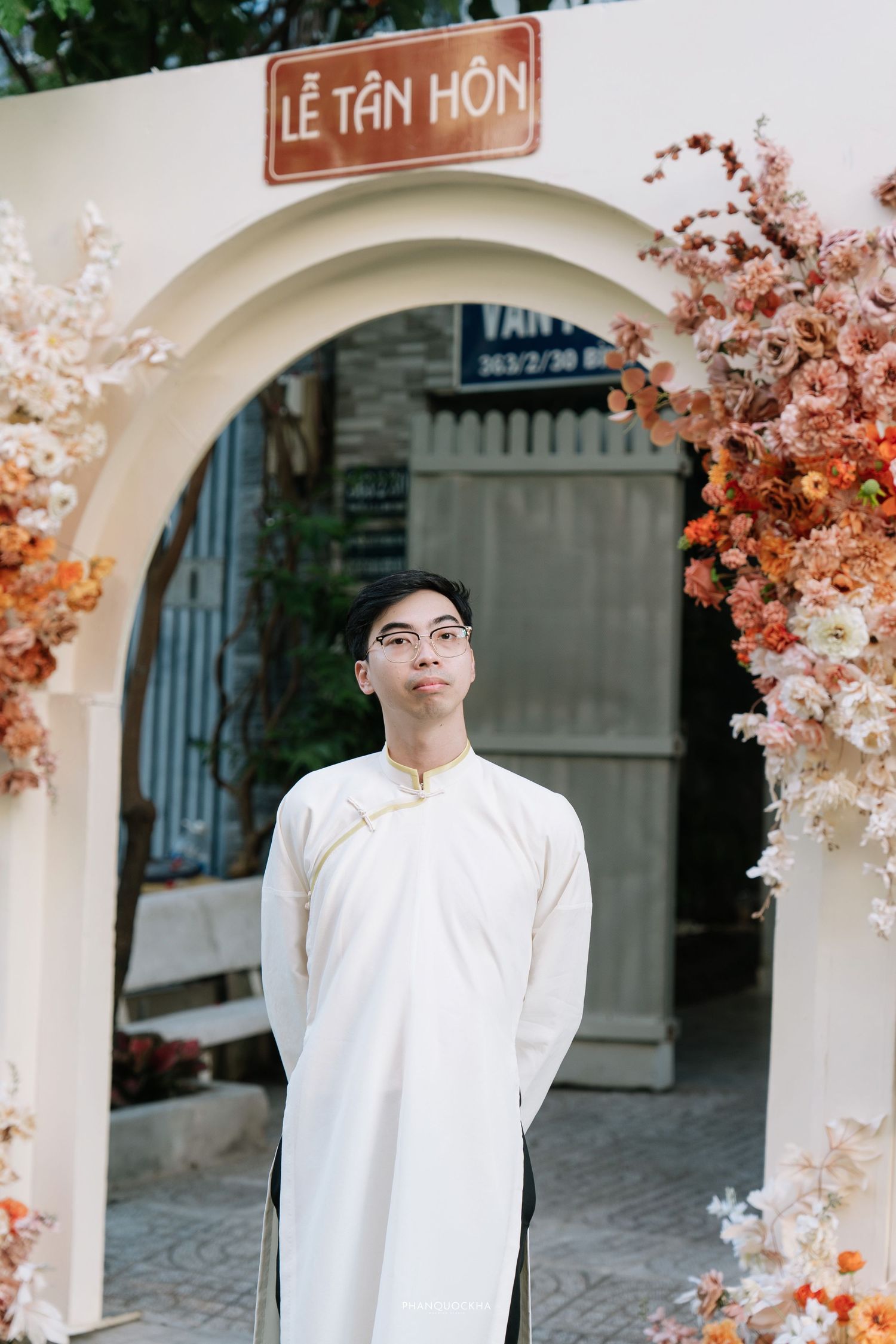 Vietnamese groom in white ao dai standing under Le Tan Hon arch decorated with autumn-toned flowers.
