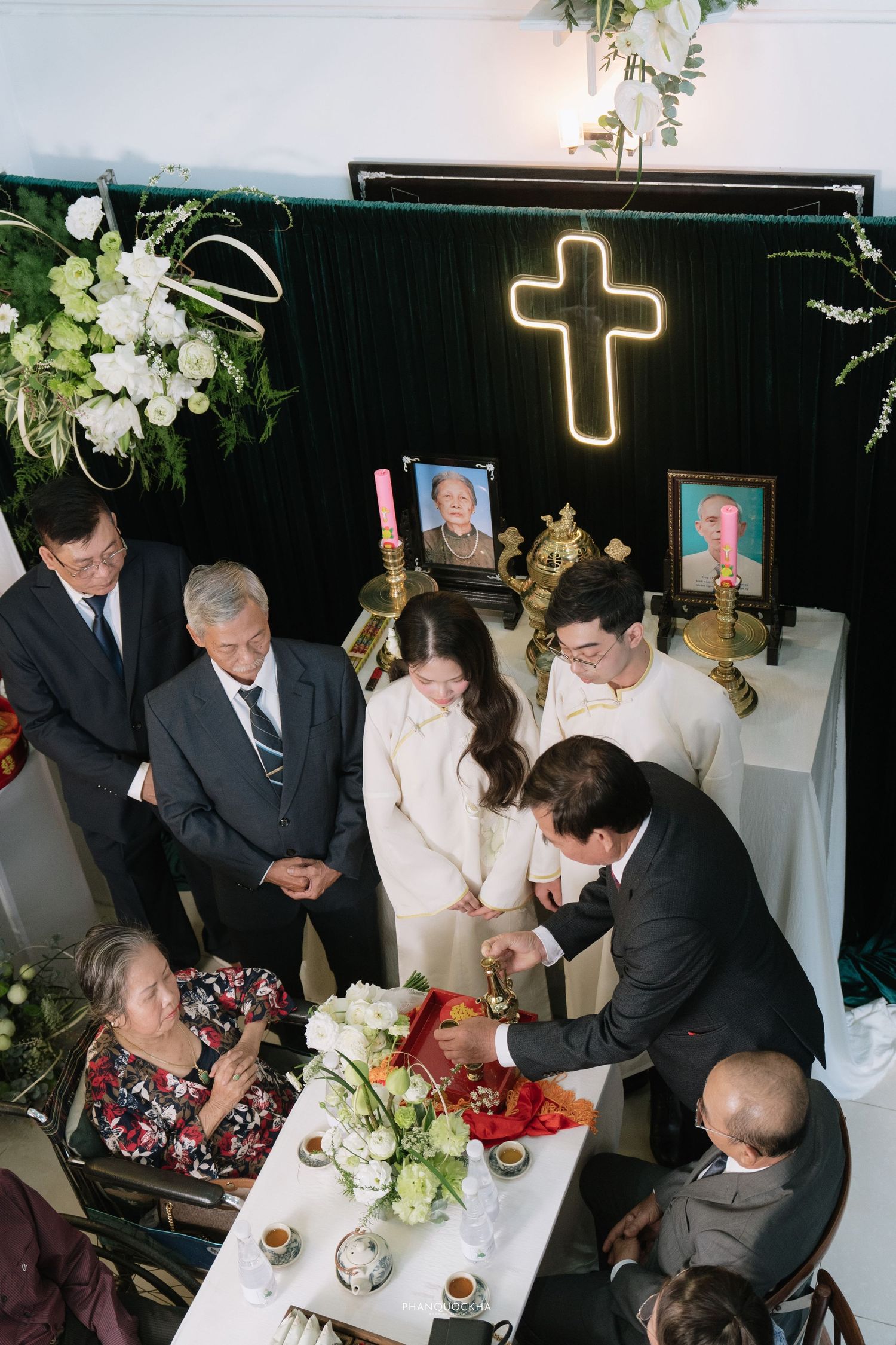 Vietnamese engagement ceremony with couple in white ao dai receiving blessings at ancestral altar with cross.