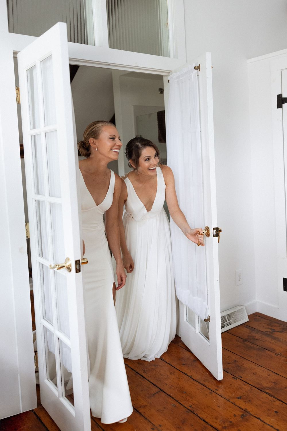 Two people in white wedding dresses sharing joyful moment through French doors.
