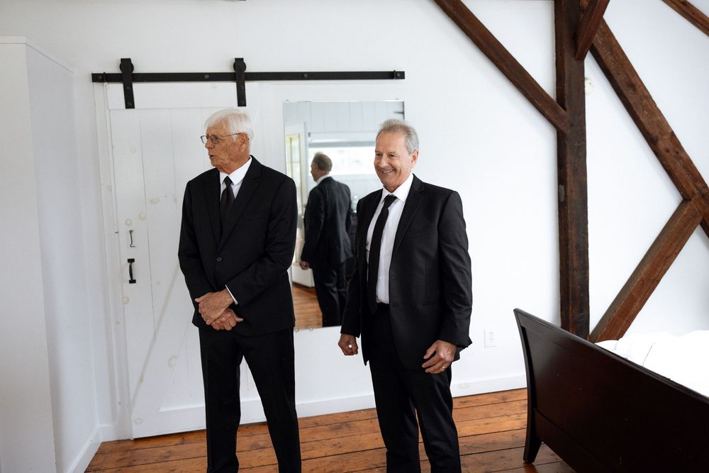 Two men in formal black suits standing in rustic room with wooden beams.
