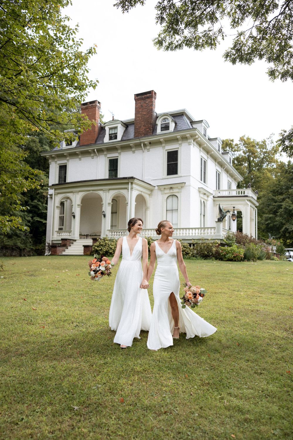 Two brides in white dresses holding bouquets pose in front of historic white mansion.