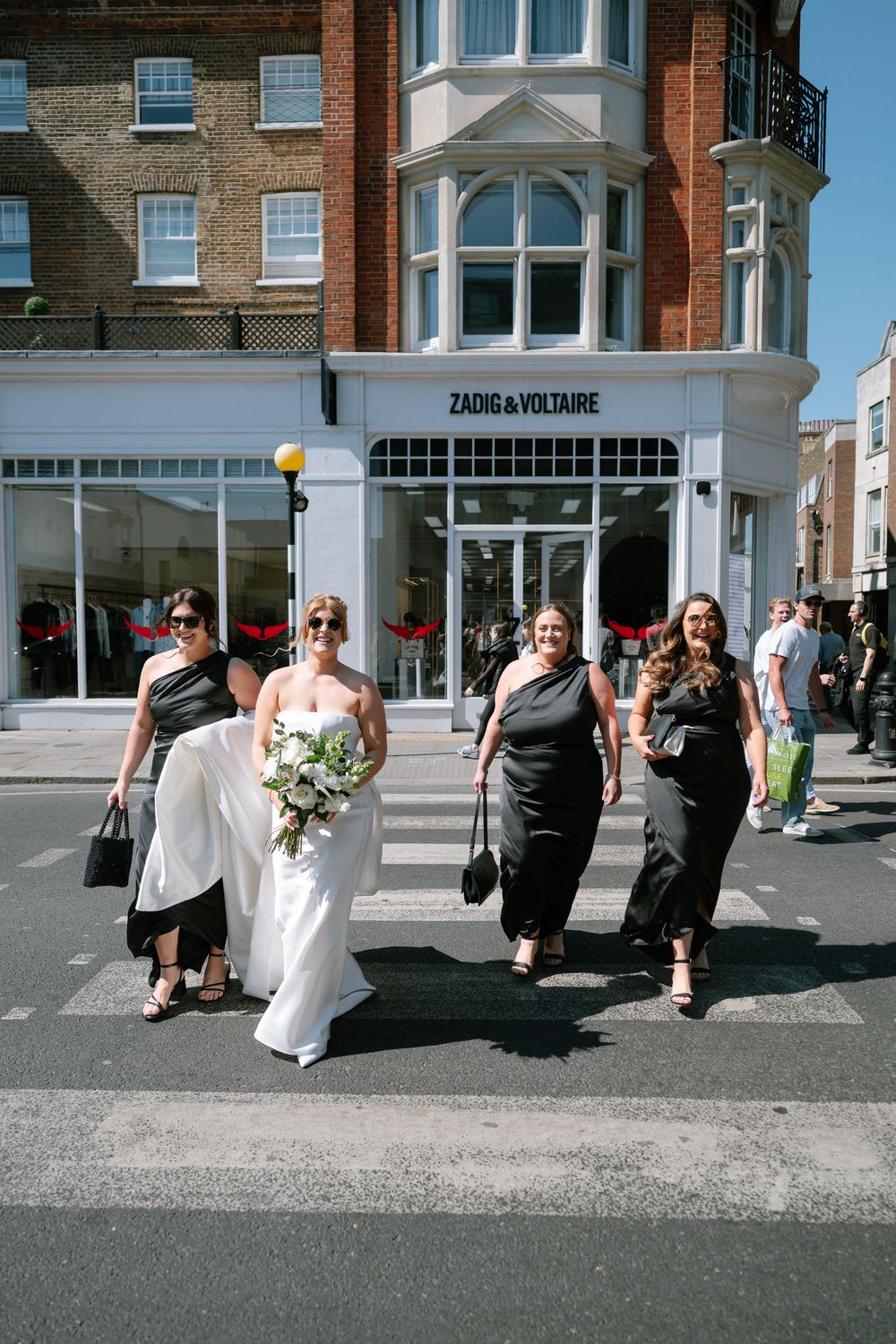 Wedding party walks down a sunny urban street past brick buildings with white architectural details.