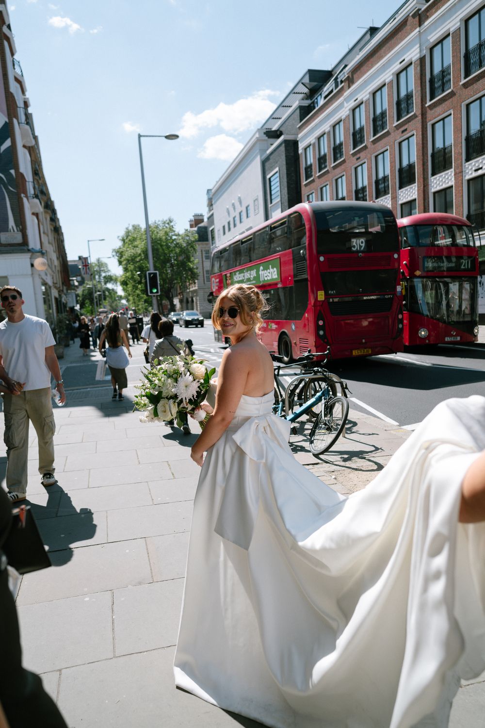 Bride in white dress walks through city street with red buses and modern buildings in background.
