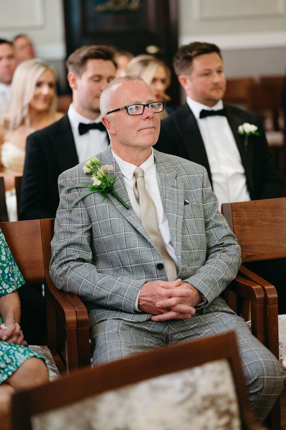 Two men wearing matching gray plaid suits and white ties sit attentively in wooden church pews during a ceremony.