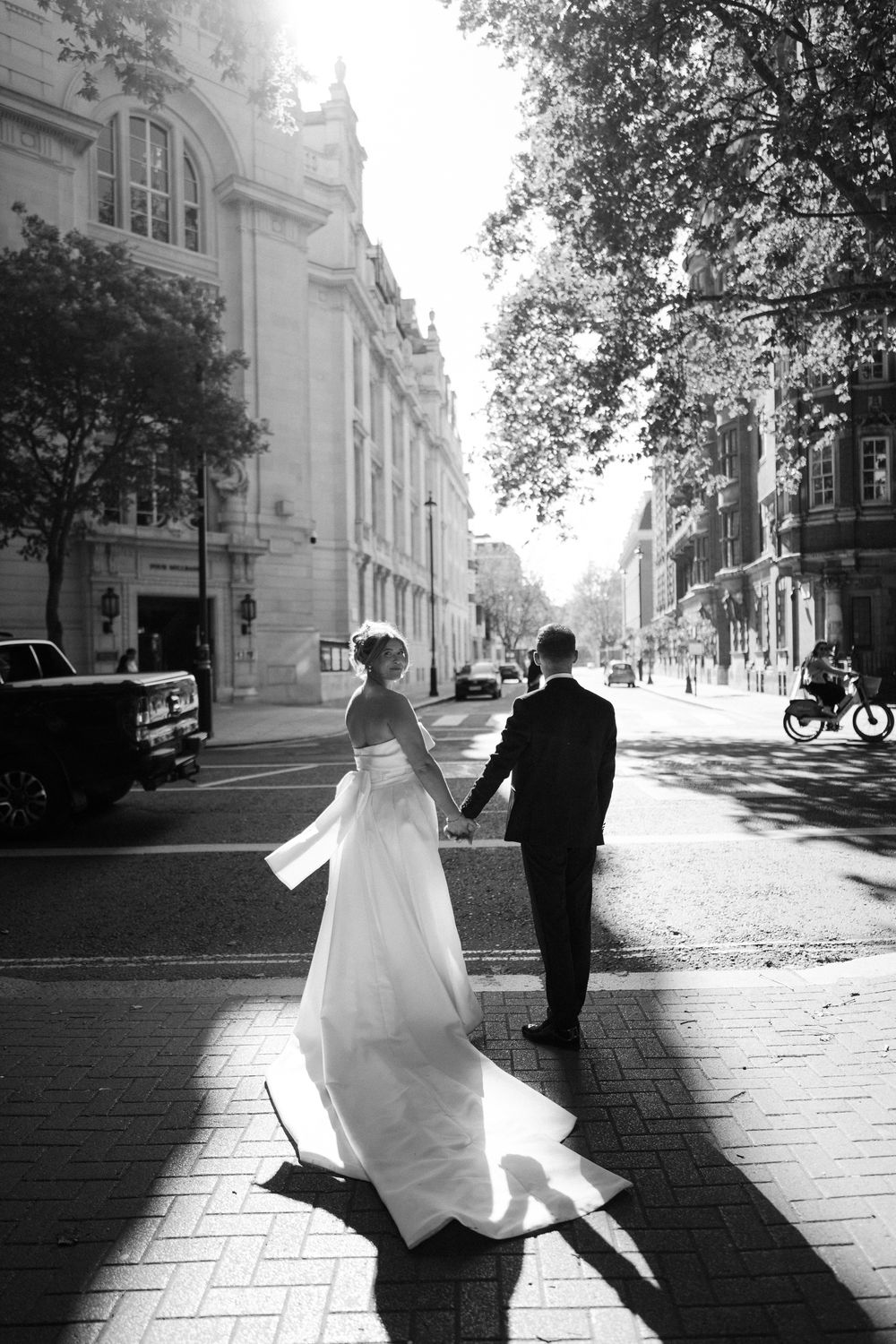 A romantic black and white series of a newlywed couple captured walking along a sunlit brick pathway in front of historic buildings.