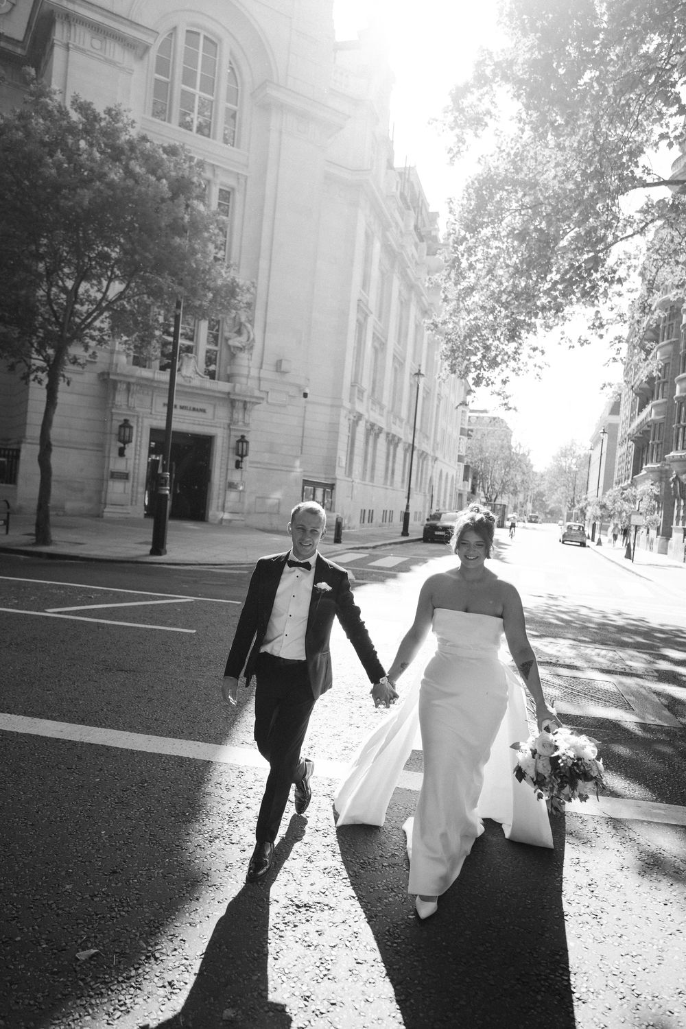 A couple walking down a bright sunlit city street while holding hands, captured in black and white photography.