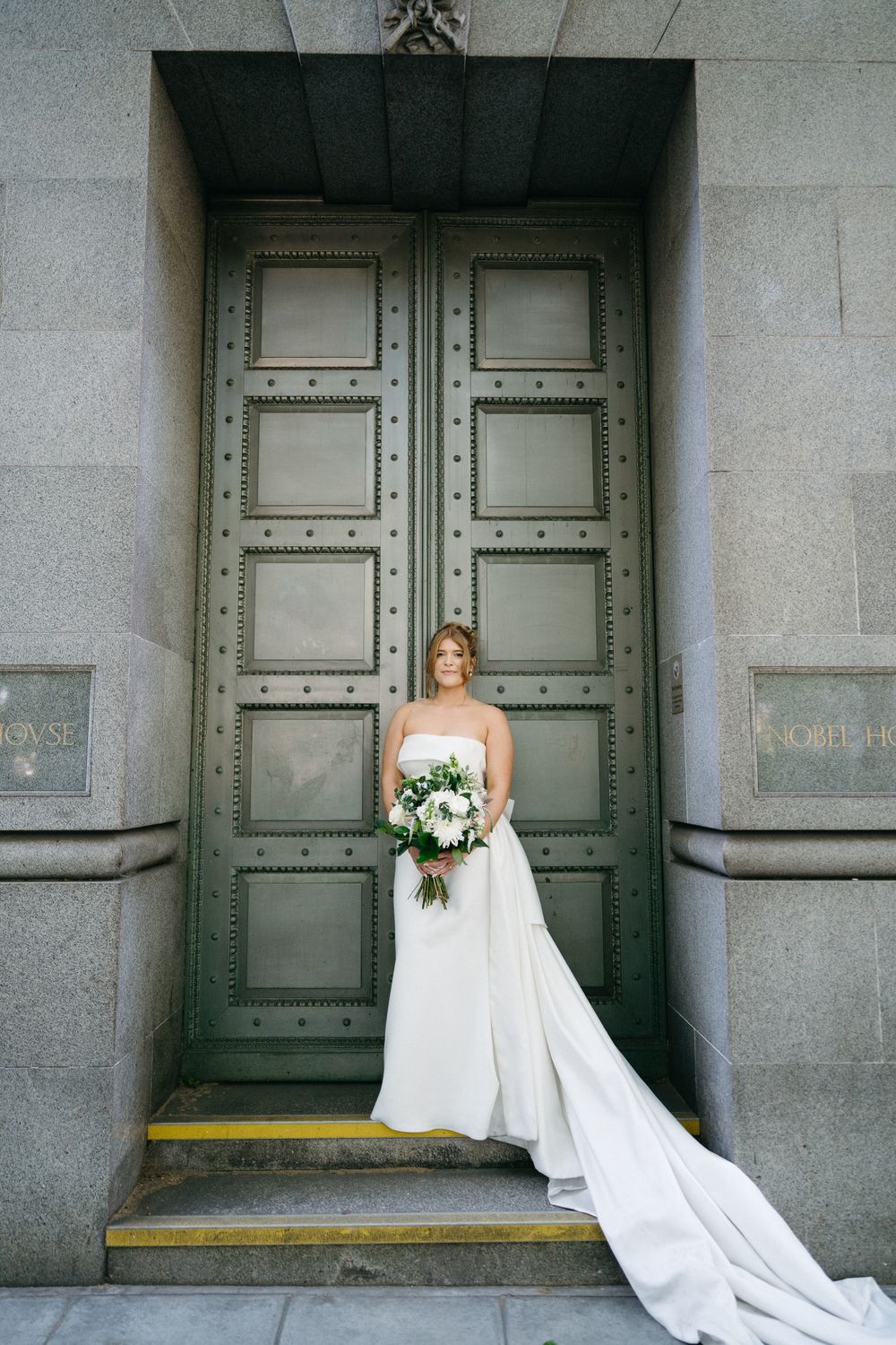 A bride in a flowing white wedding gown with long train poses in front of grand metallic doors in stone archway.