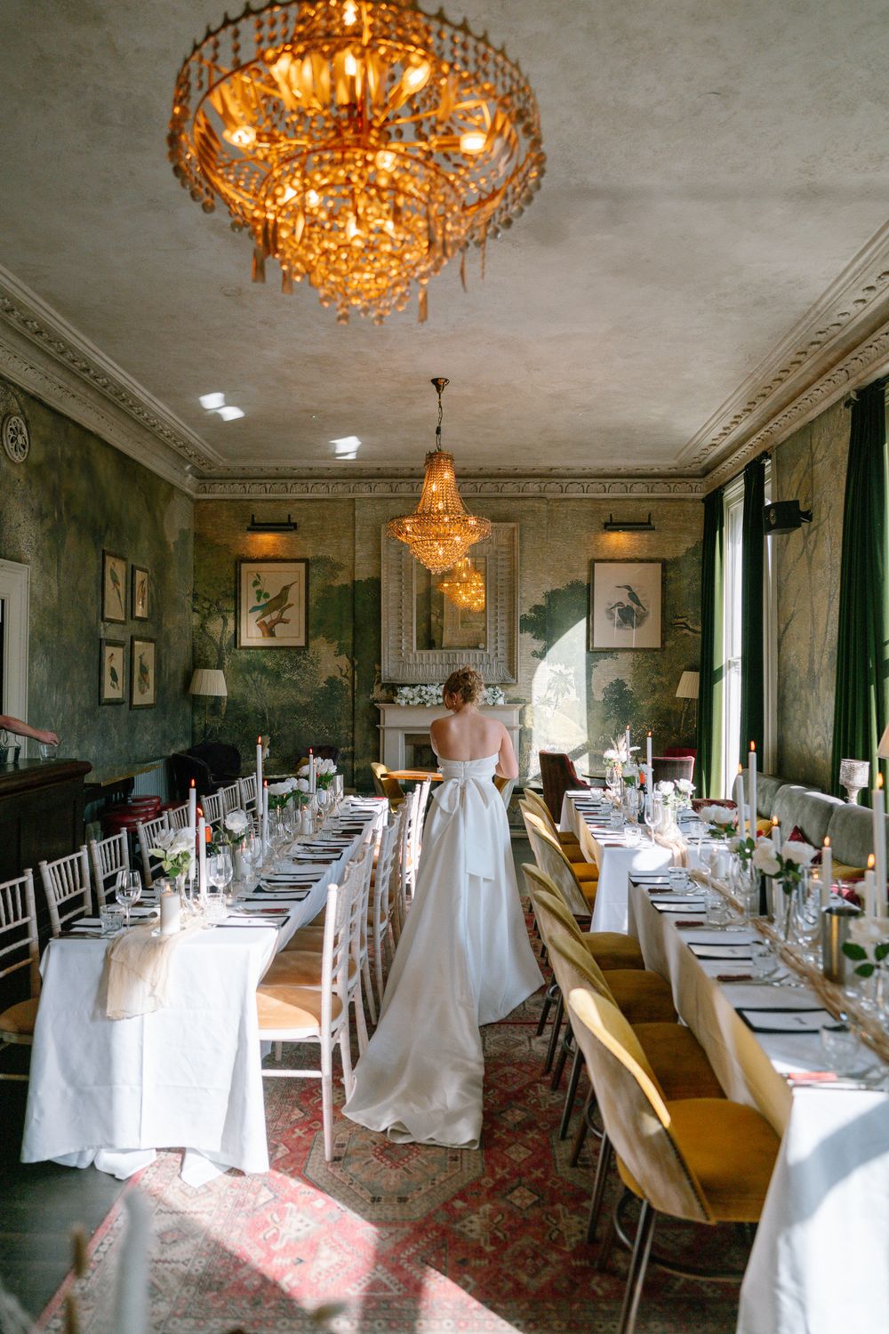 Elegant dining room with ornate crystal chandeliers, white-clothed tables, and green wallpaper in a vintage wedding venue setting.