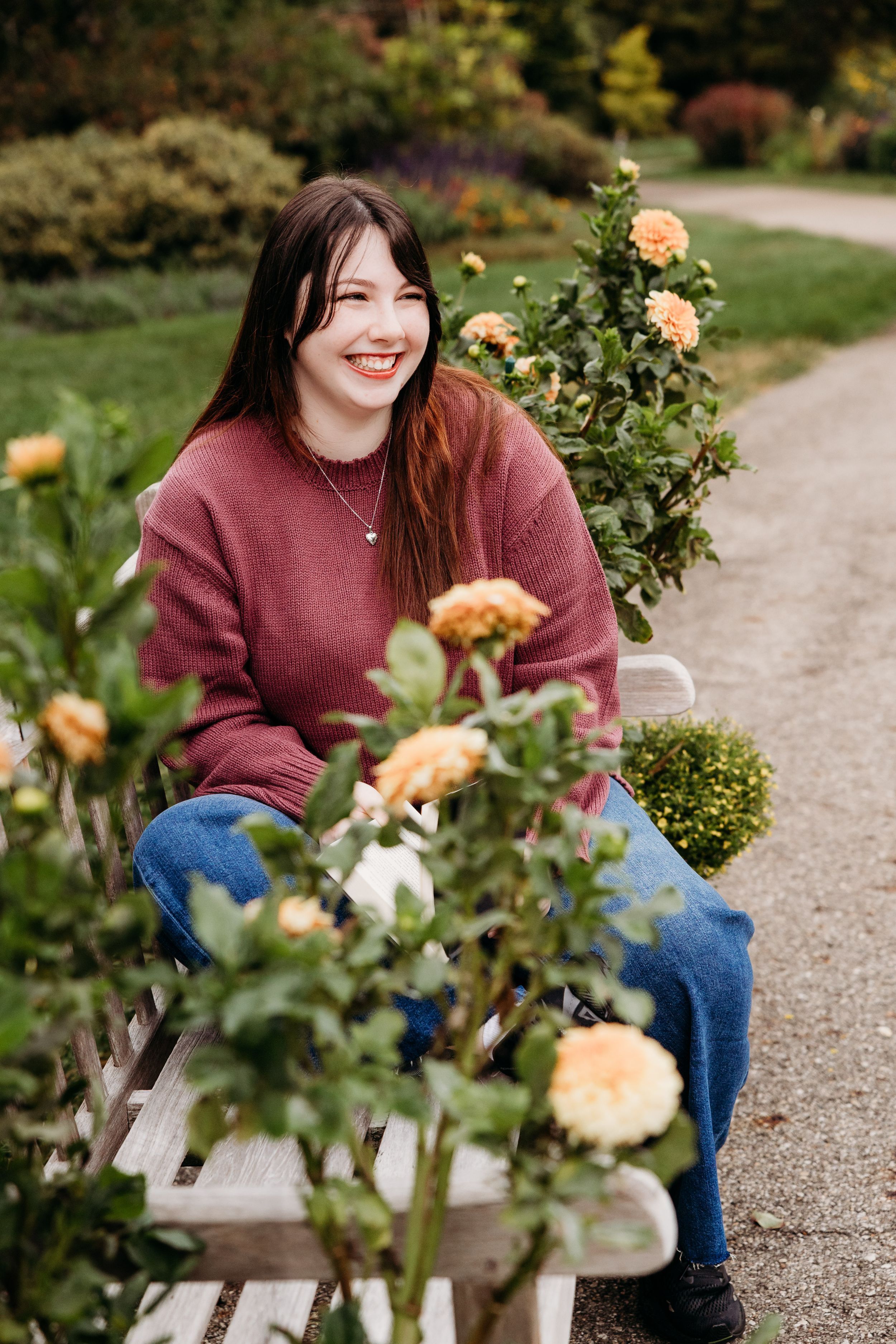 A person in a burgundy sweater and jeans sits on a garden bench surrounded by blooming peach roses.