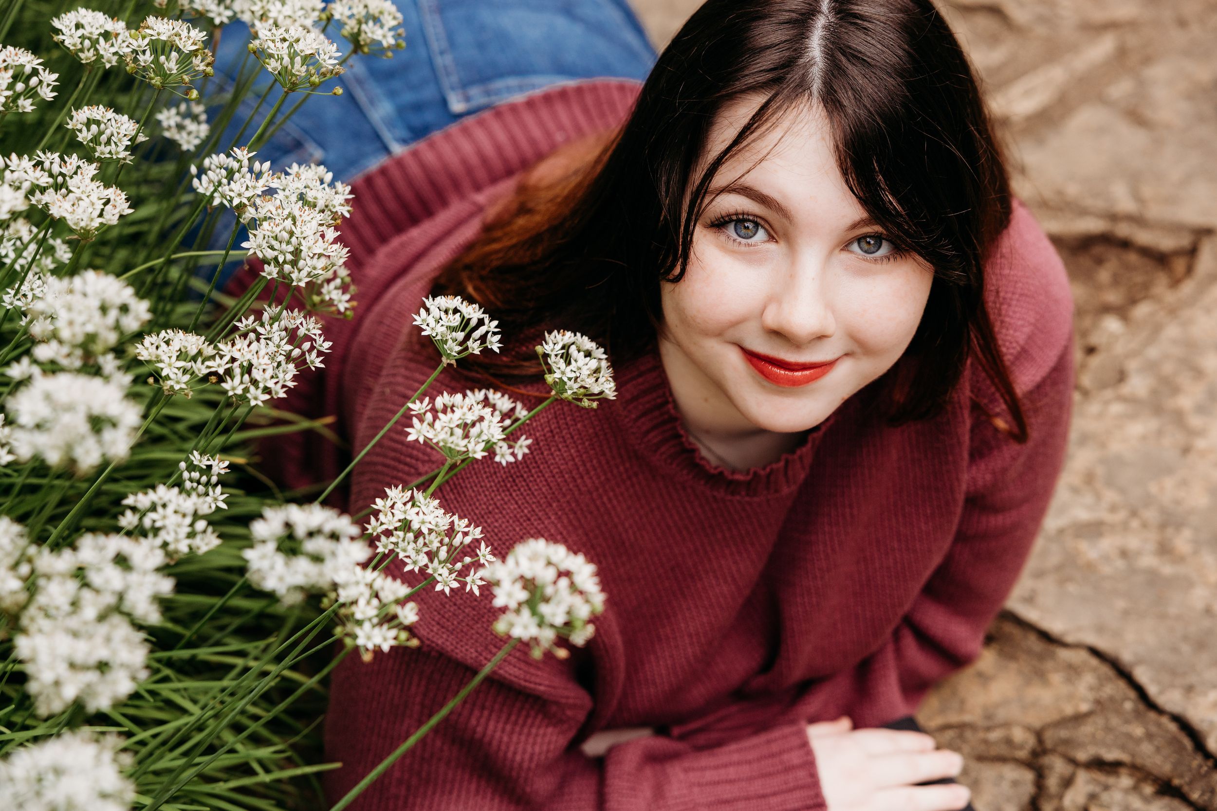 A woman in a burgundy sweater poses among delicate white flowers and lush greenery against a rustic stone background.