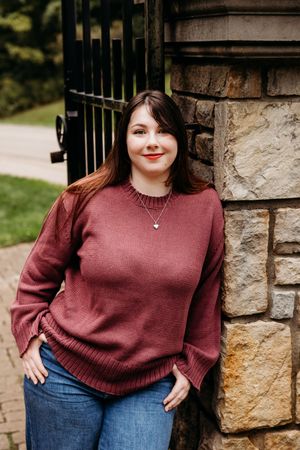 Person in burgundy sweater and jeans poses next to stone wall and iron gate outdoors.