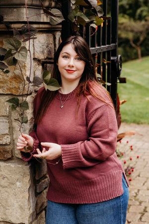 A person in a burgundy sweater and jeans poses by a stone wall with autumn foliage.