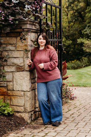 A person in a burgundy sweater and denim skirt stands near a rustic stone pillar on a paved garden pathway in autumn.