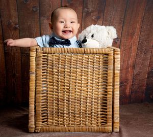 A smiling baby peeks out of a wicker basket against a rustic wooden backdrop with a heartwarming expression.