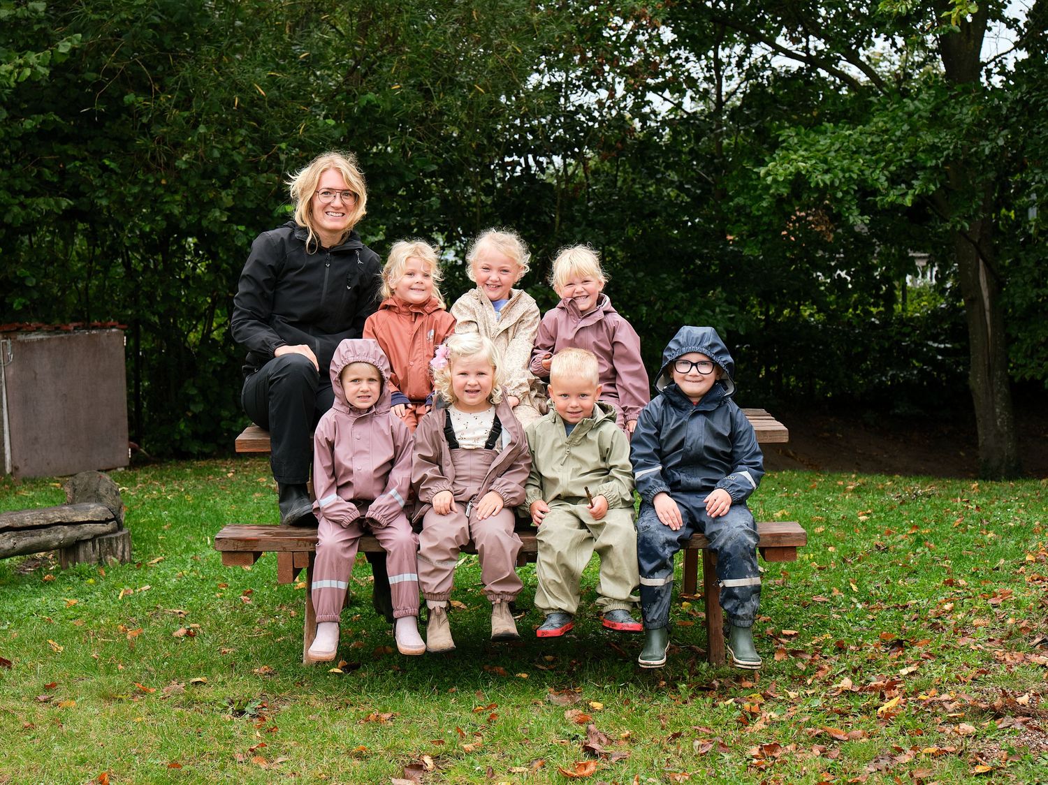Children in winter coats sitting together on outdoor steps and benches surrounded by autumn leaves.
