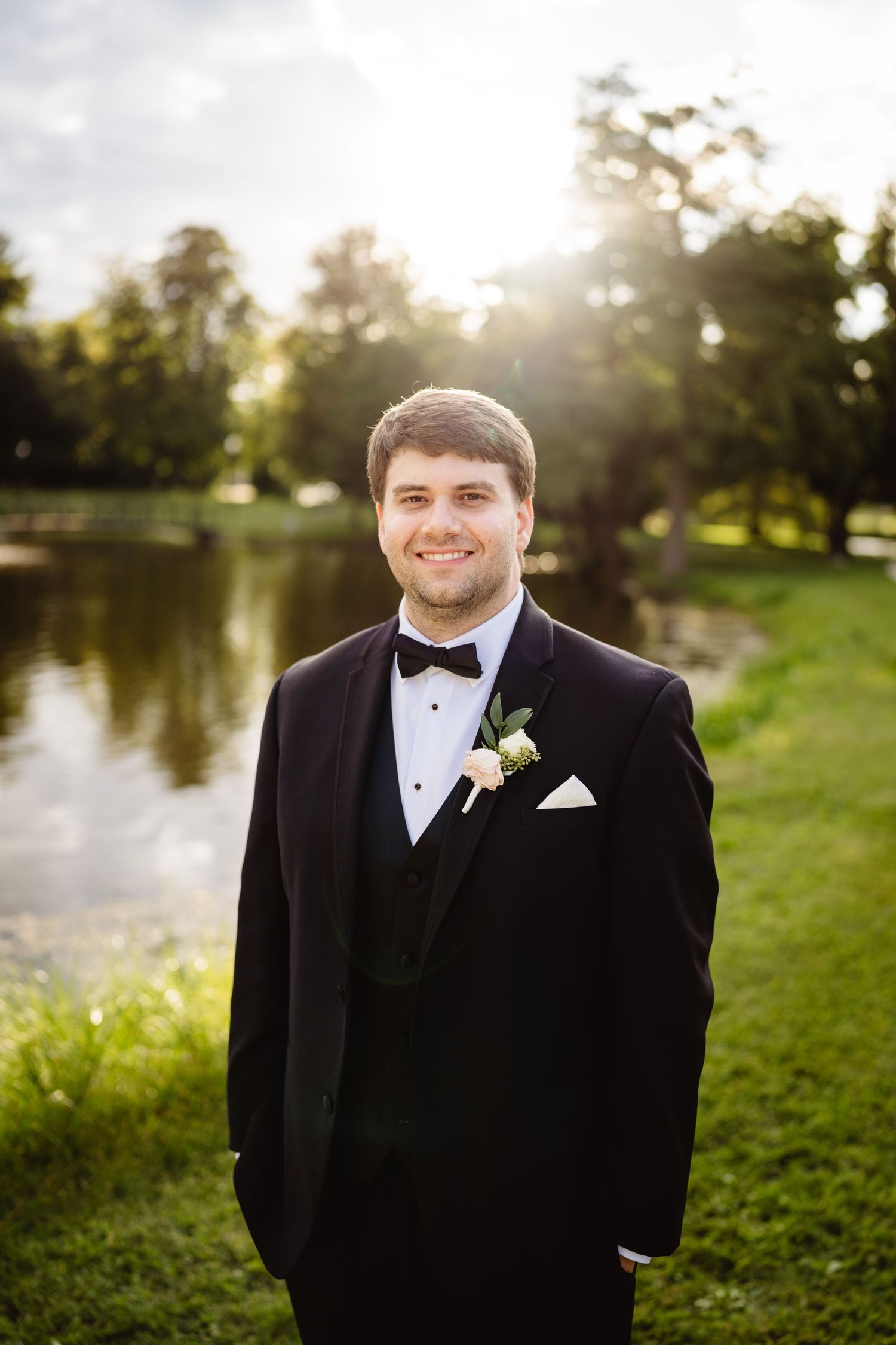 Groom in classic black tuxedo stands by lakeside during sunset on wedding day.