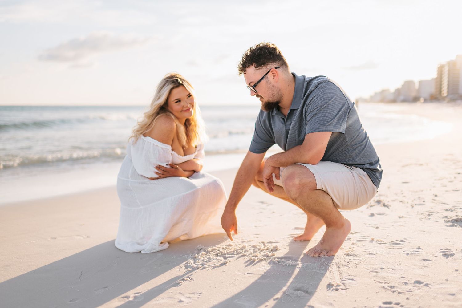 Playful couple poses on white sandy beach with ocean and cityscape in background during sunset.
