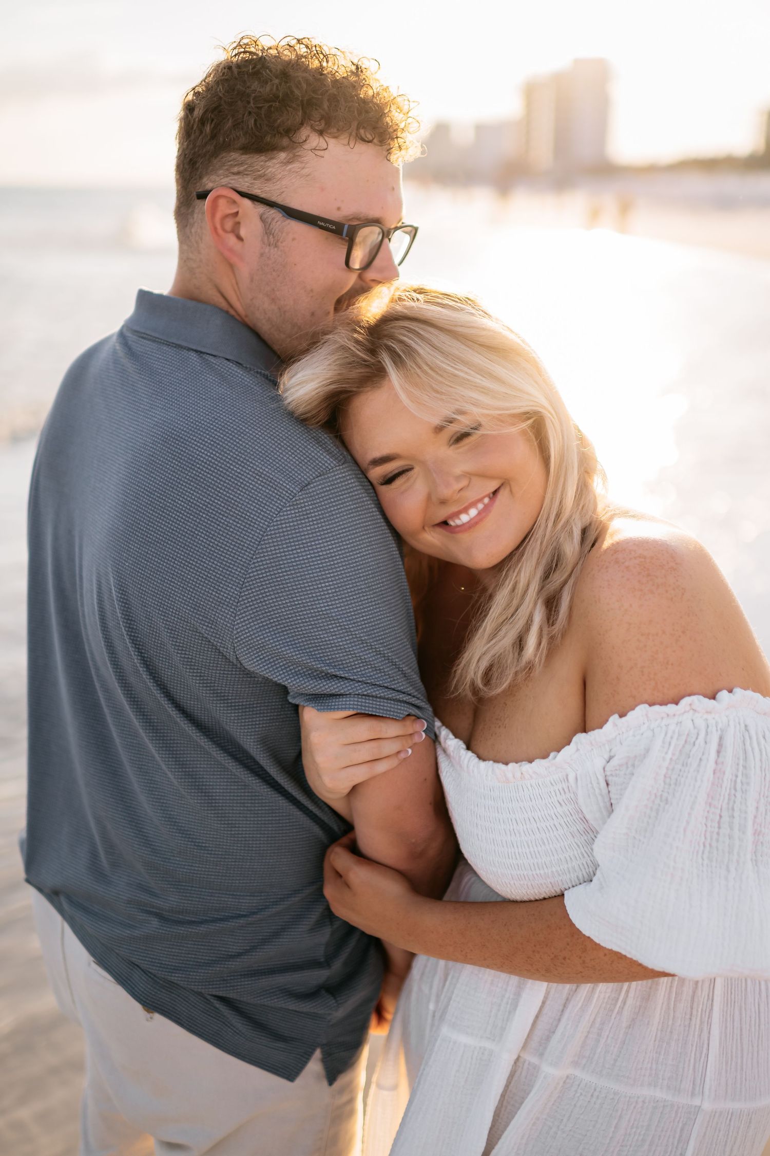 A romantic couple embraces on a sunlit beach wearing white and grey casual attire during a photo session.