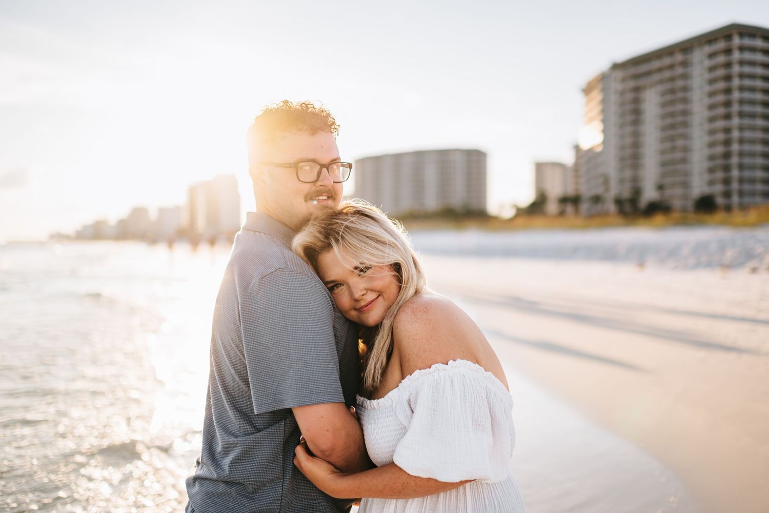 Couple enjoys tender moment on beach with coastal high-rises and setting sun creating warm ambiance.