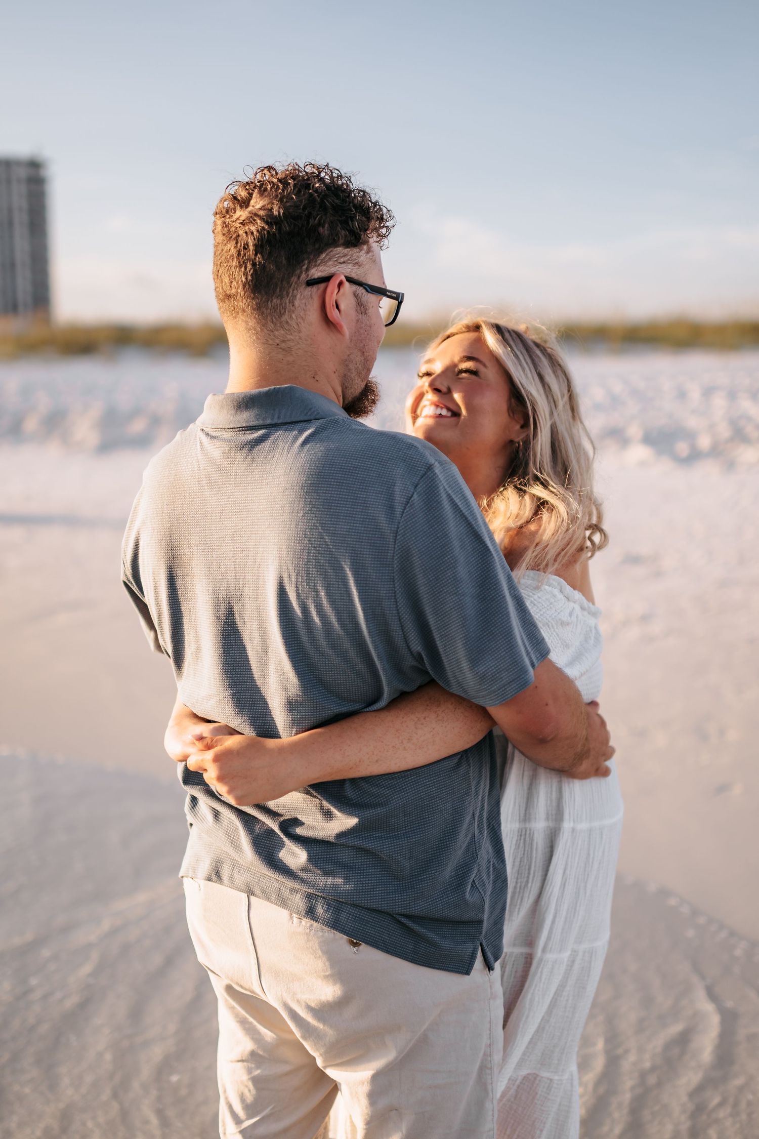 A couple embraces on a white sandy beach at sunset wearing casual grey and white clothing.