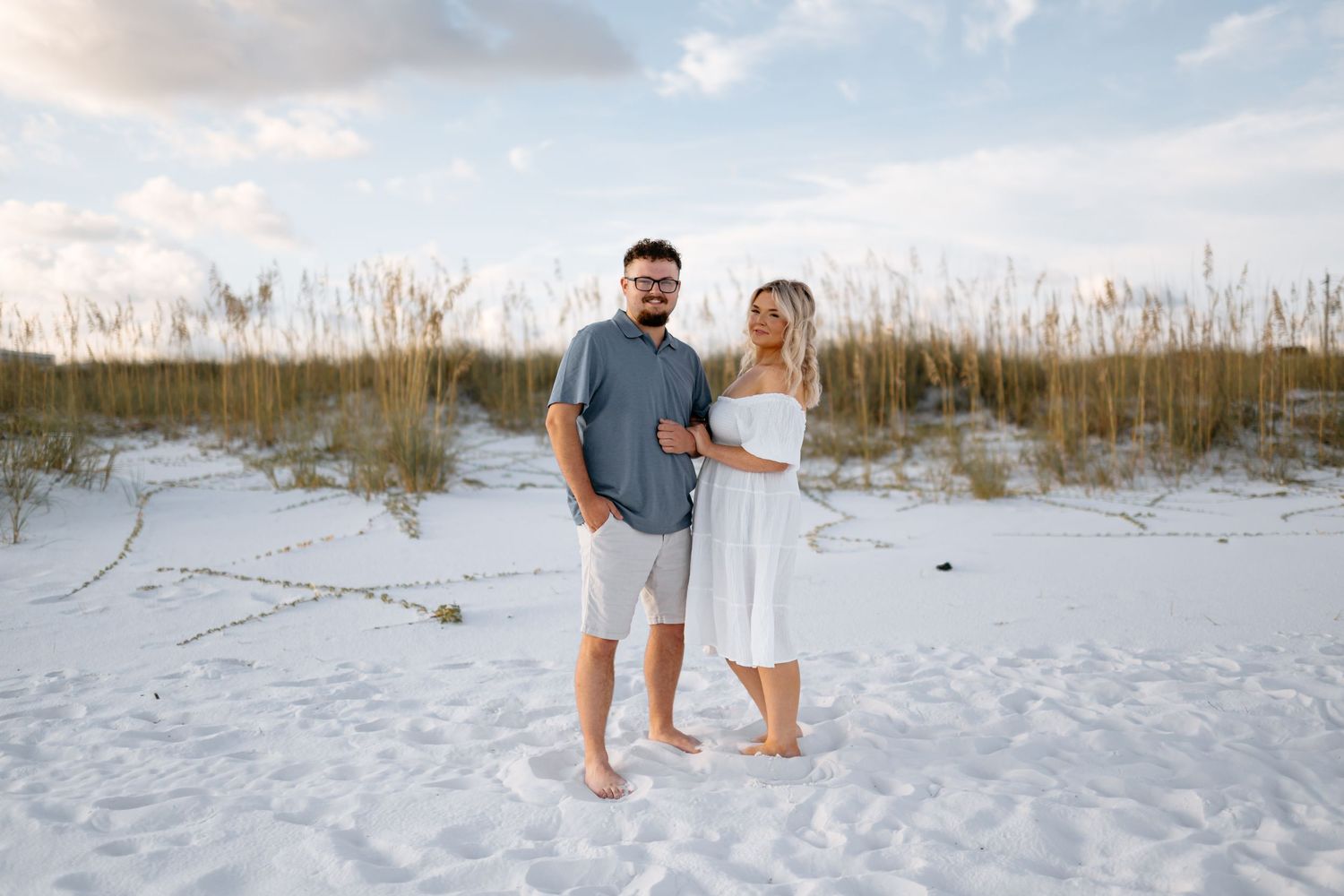 Couple walking barefoot along white sandy beach with sea oats and dunes in the background at sunset.