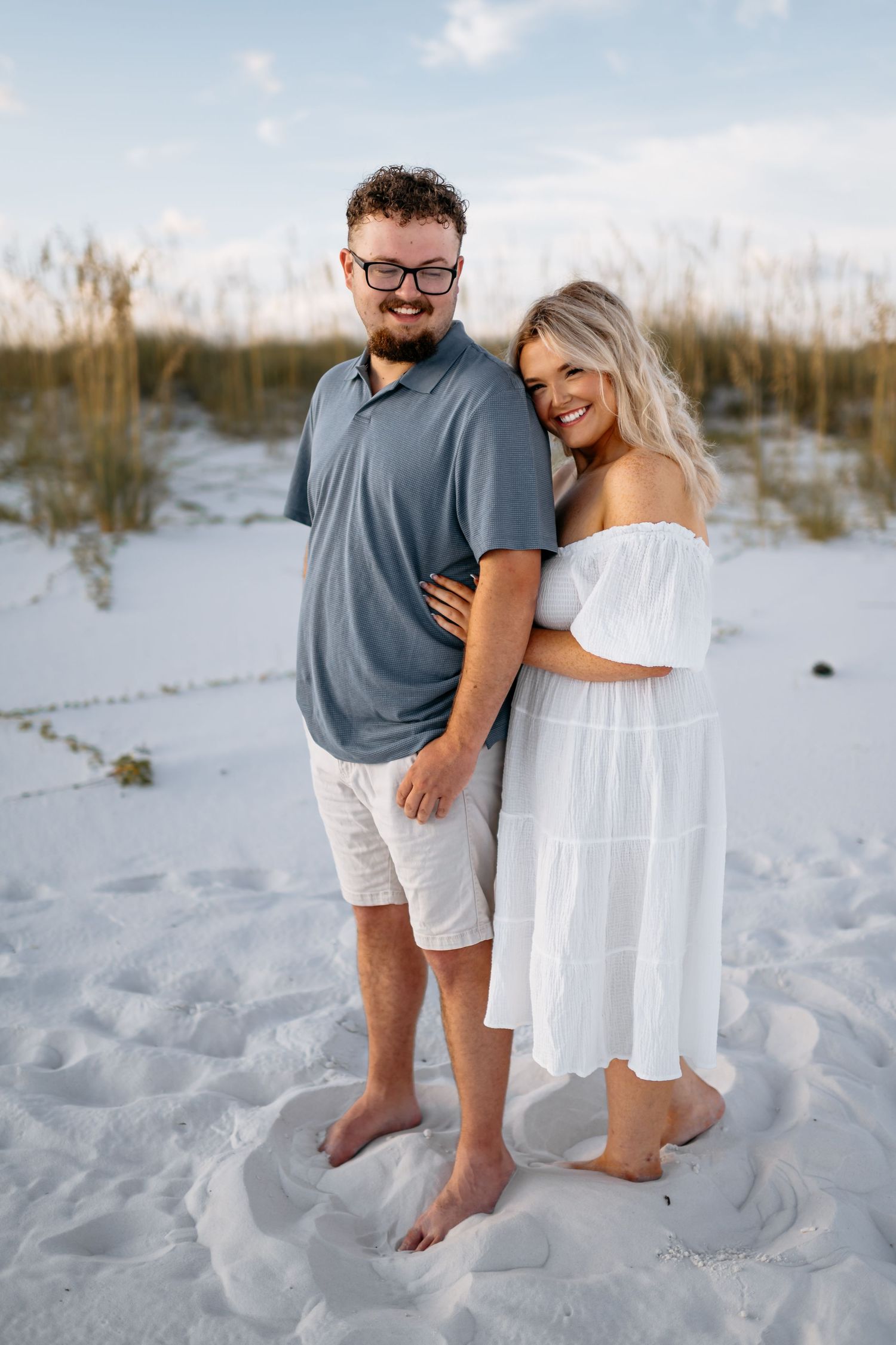 A couple shares tender moments together on a white sandy beach with sea oats in the background during sunset.