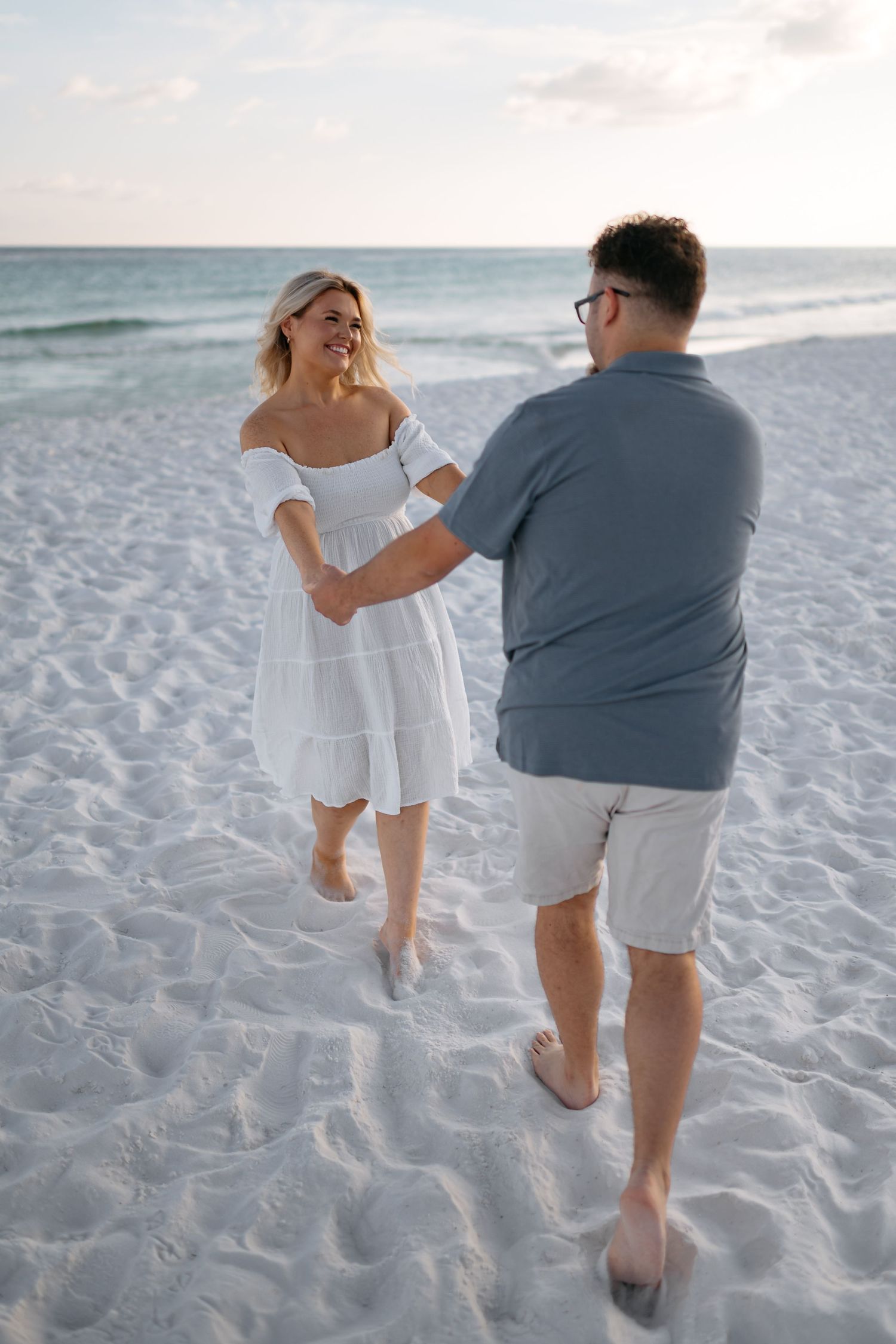 A couple walking together on a white sandy beach at sunset with the ocean in the background.
