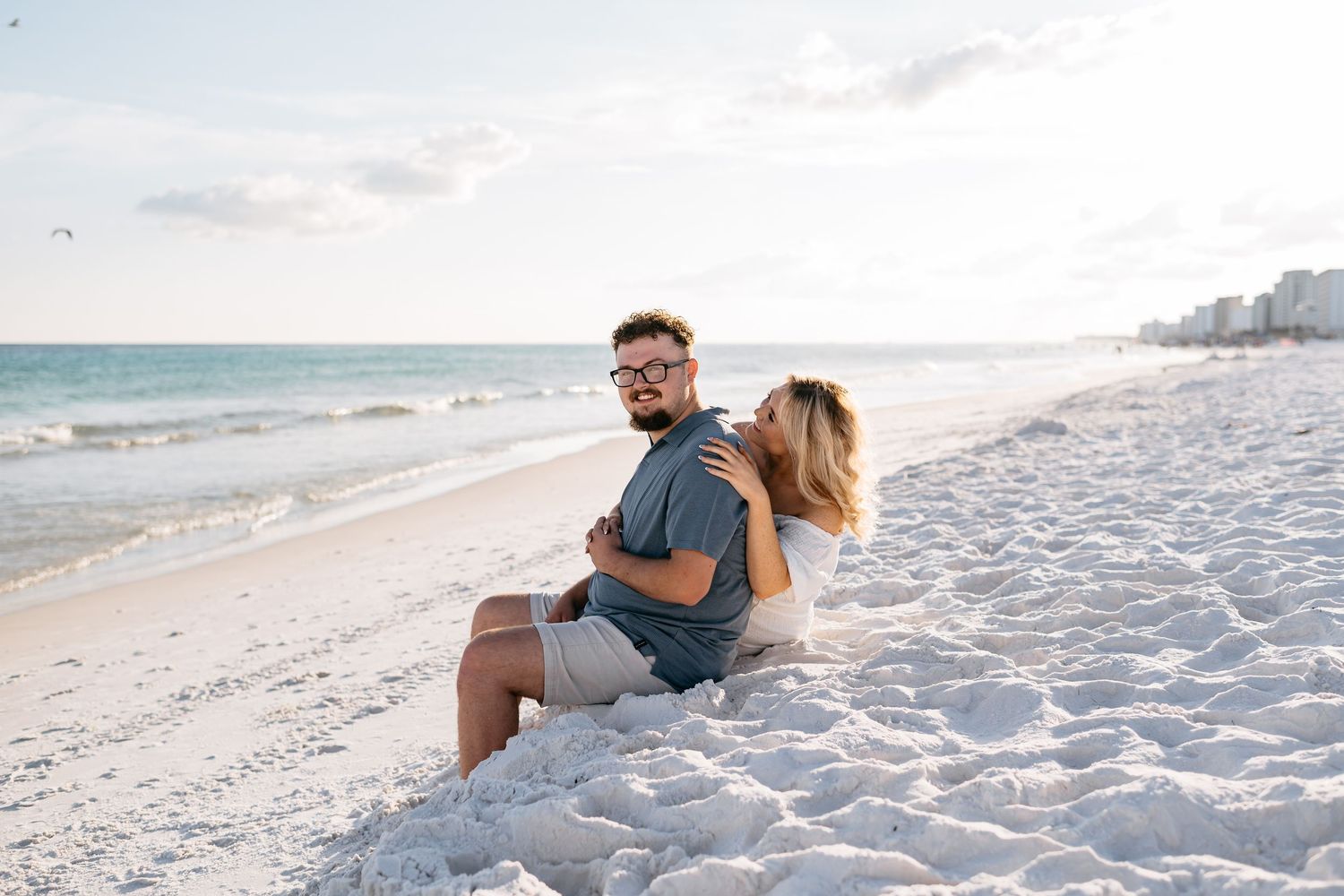 A couple enjoys a tender moment while sitting on a sandy beach with waves and coastline in the background.