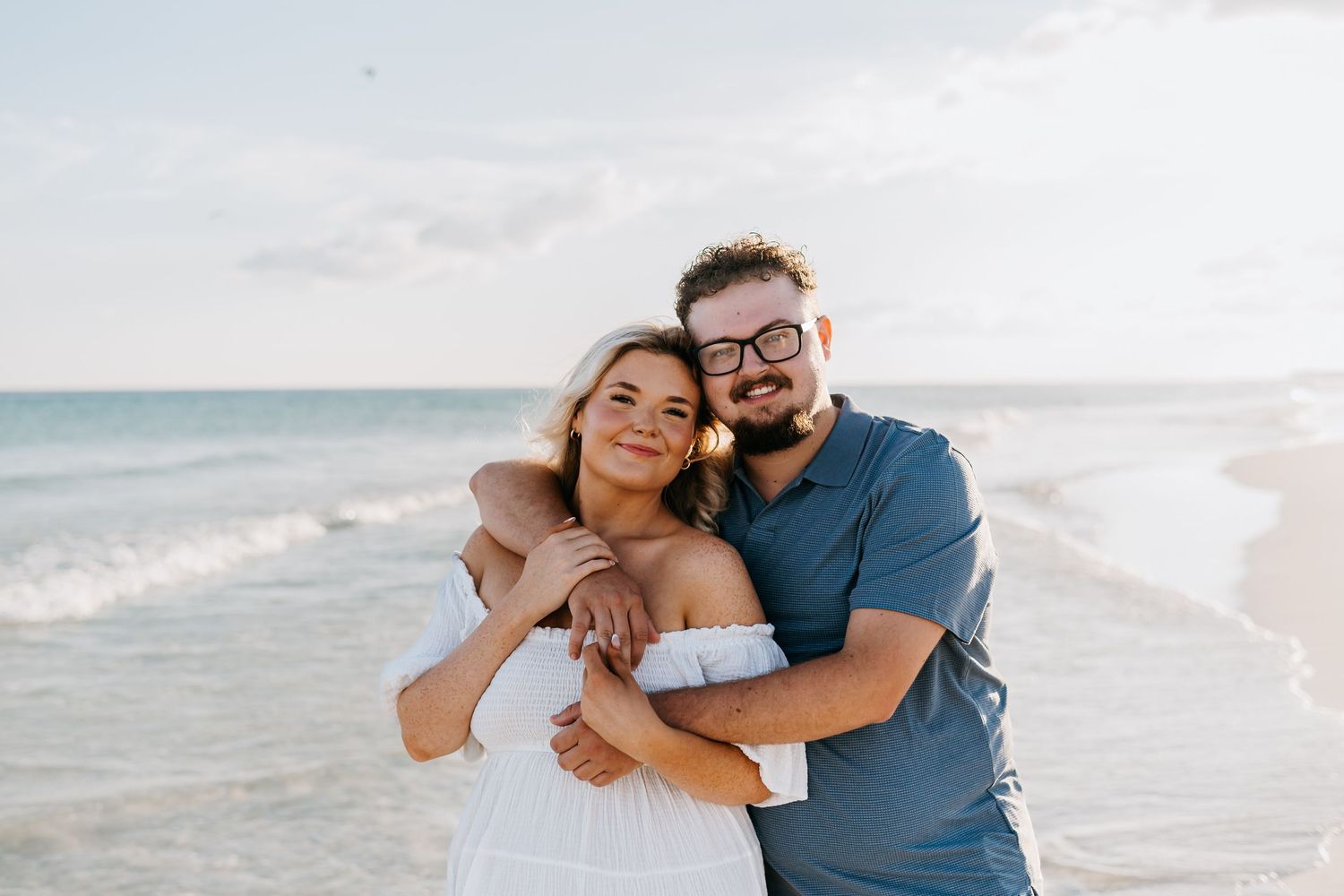 A happy couple embracing on the beach with the ocean and cloudy sky behind them.