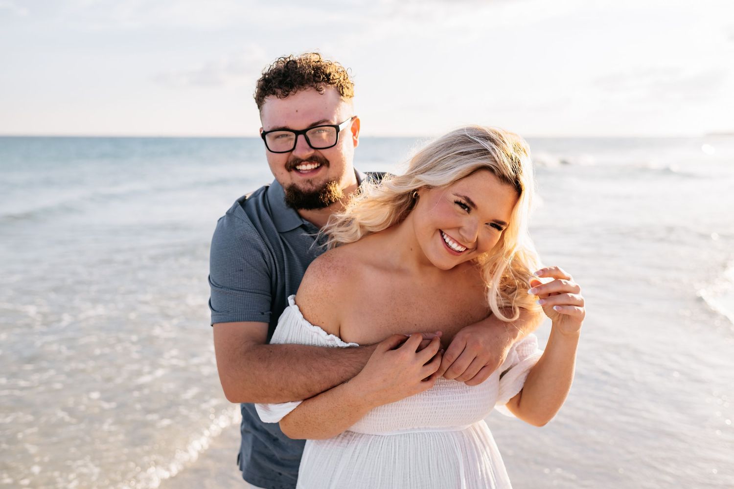 Romantic couple embraces on sandy beach during golden hour with ocean waves and high-rise buildings in background.